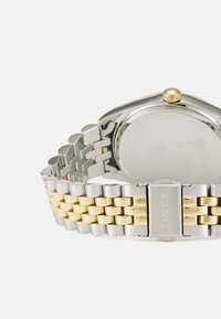 Montre Timex avec un boîtier en acier inoxydable, un bracelet en métal doré et argenté, un cadran rond et une couronne texturée dorée.
