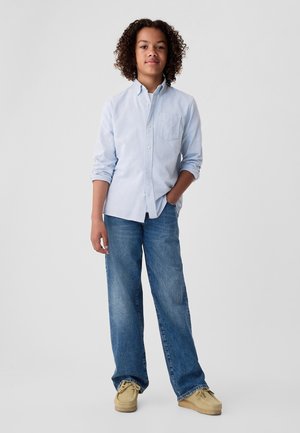 Lichtblauw overhemd met lange mouwen en een zak, gecombineerd met een blauwe spijkerbroek en beige mocassin schoenen. Effen achtergrond, frontale aanzicht.