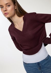 Burgundowa sweter z dzianiny z dekoltem w serek i ściągaczem u dołu, zestawiony z białą koszulką i niebieskimi dżinsami. Sweter ma luźny krój.