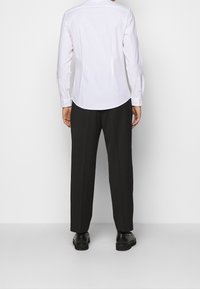 Weißes Langarm-Hemd mit Knopfleiste; einfarbiger Stoff; abgerundeter Saum; kombiniert mit schwarzen Hosen und schwarzen Schuhen; klassischer Schnitt, minimalistisches Design.