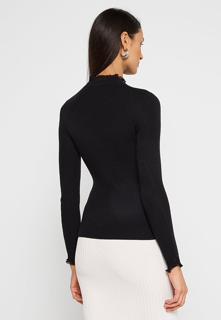 Kobieta ubrana w czarną, długą bluzkę z prążkowanej tkaniny oraz białą spódnicę z prążkowanej tkaniny, stojąca na jednolitym jasnym tle, zwrócona plecami.