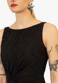 Schwarzes Kleid aus strukturiertem Stoff mit subtiler Schimmeroptik. Asymmetrischer Ausschnitt und gedrehte Detail an der Taille. Silberne Statement-Ohrringe.