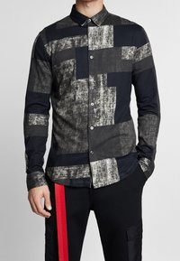 Chemise à manches longues avec boutons, présentant un motif géométrique en noir, gris et blanc. Fabriquée dans un tissu doux avec une texture lisse.