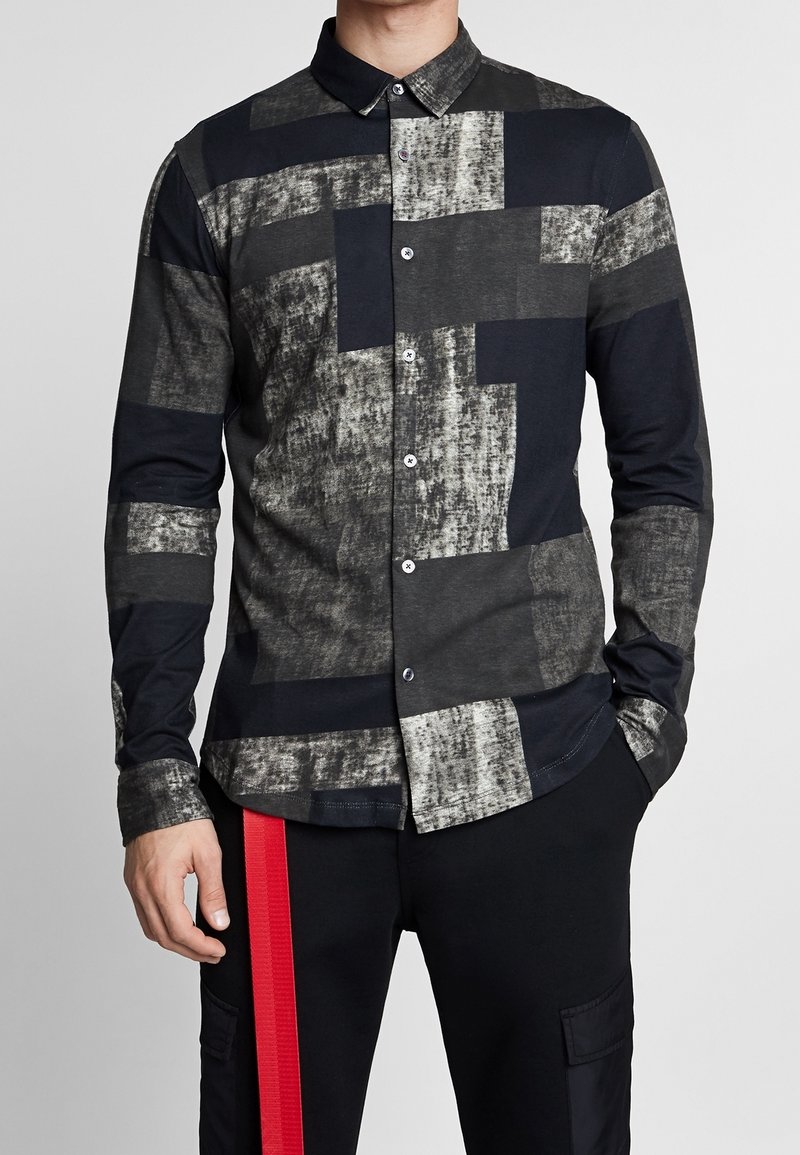 Chemise à manches longues avec boutons, présentant un motif géométrique en noir, gris et blanc. Fabriquée dans un tissu doux avec une texture lisse.
