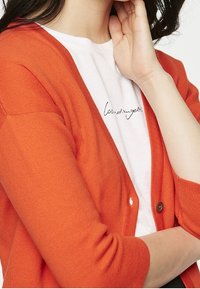 Orange stickad kofta med trekvartsärmar, rund hals och knappstängning över en vit t-shirt med kursiv text.