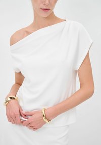 Top bianco off-shoulder realizzato in tessuto morbido con maniche corte. Il modello indossa due bracciali ondulati dorati ai polsi.