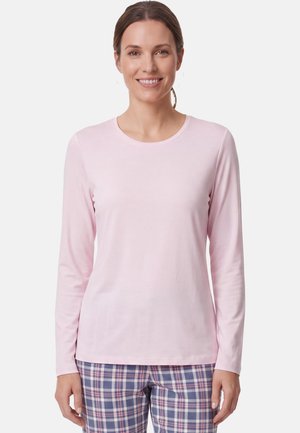 Lächelnde Frau mit braunen Haaren im Pferdeschwanz, die ein hellrosa Langarmshirt und blau-rosa-weiße karierte Hose trägt.