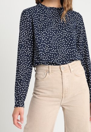 Mujer con blusa de manga larga azul marino con lunares blancos y pantalones beige de cintura alta, de pie con un brazo relajado.