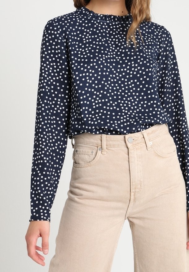 Femme portant un chemisier bleu marine à manches longues avec des pois blancs et un pantalon beige taille haute, debout avec un bras détendu.