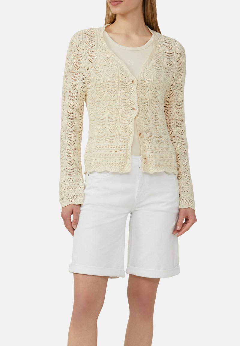 Cremfarbener Cardigan aus offener Strickware mit gewelltem Saum, langen Ärmeln und Knopfverschluss, kombiniert mit einem beigen Top und weißen Shorts.