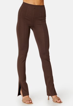 Pantalon marron ajusté avec coutures devant et fentes aux chevilles porté avec des sandales beige tressées à talons hauts sur un fond clair.