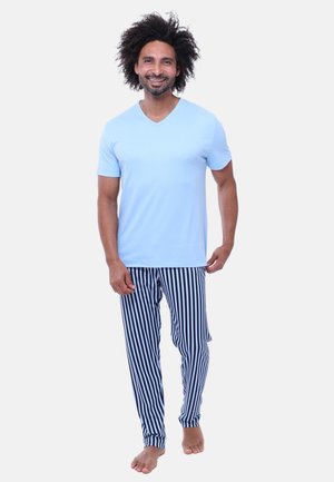 Lachende man met krullend haar, gekleed in een lichtblauw shirt met korte mouwen en blauw-witte verticale gestreepte broek, blootsvoets, staand op een witte achtergrond.