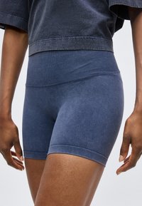 Gros plan d'une personne portant un short de sport bleu taille haute et un top court gris foncé, debout avec les bras détendus devant un fond uni.