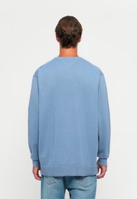 Uomo con capelli corti e castani, che indossa un maglione blu e jeans azzurri, in piedi di spalle su uno sfondo bianco neutro.