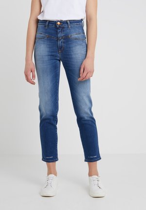 Jeans de corte ajustado con degradado azul, usados por una persona de pie, combinados con zapatillas blancas y una camiseta blanca metida por dentro.