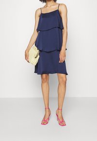 Mörkblå ärmlös klänning med lagerdesign, mjuk textur och utsvängd nederkant. Matchad med en ljusgul vävd clutch och rosa sandaler.