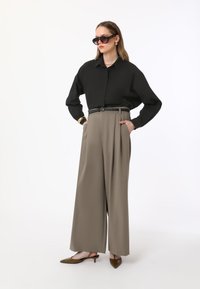 Black oversized button-up shirt, taupe pleated high-waisted trousers, and brown pointed-toe slingback heels. Minimalist gold accessories.