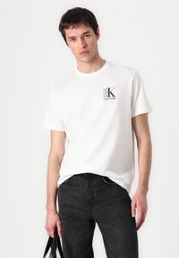 T-shirt en coton blanc avec un design de logo carré en gris et bleu de Calvin Klein, assorti à un jean noir.