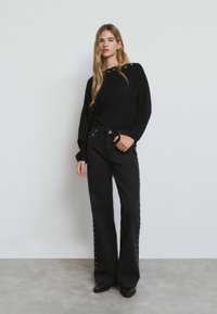 Chemise noire à manches longues avec des accents de boutons aux épaules, associée à un jean noir à large jambe avec des fermetures à pression sur les côtés.