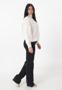 Witte oversized gebreide trui met een hoge nek, gedragen met zwarte flared jeans en doorzichtige haksandalen. Gladde textuur, relaxte pasvorm.