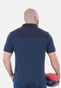 Polo shirt navy à manches courtes, avec un panneau d'épaule bleu foncé et un tissu texturé, porté par une personne tenant un ballon de basket.