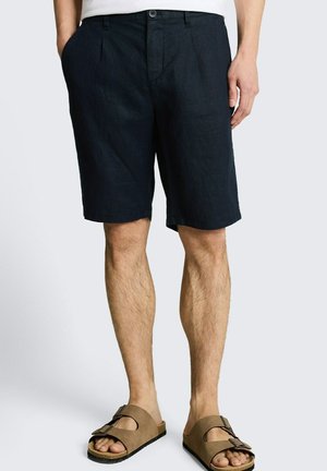 Man draagt zwarte knielange shorts en bruine sandalen met gesp, staand tegen een effen lichte achtergrond.