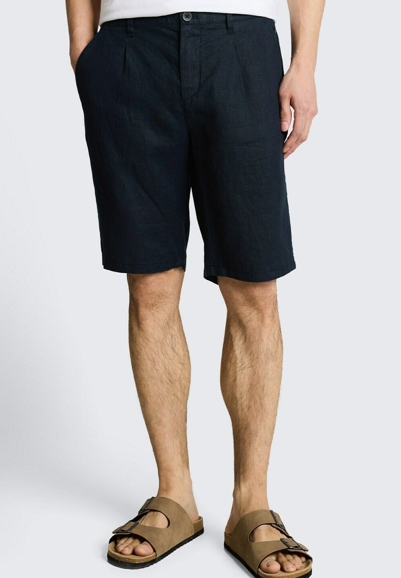 Hombre con pantalones cortos negros hasta la rodilla y sandalias marrones con hebilla, de pie contra un fondo claro y liso.