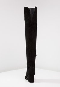 Mustat yli polven saappaat, jotka on valmistettu mokasta, neliönmuotoisella korolla, sivussa vetoketjukiinnitys ja ylhäällä V-muotoinen leikkaus.