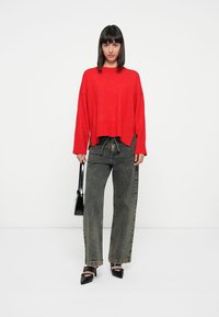 Rode cropped sweater, losse pasvorm. Olijfgroene wijde broek. Zwarte puntige hakken. Zwarte handtas. Effen witte achtergrond.