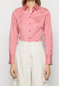 Camicia a maniche lunghe in satin rosa chiaro, con colletto a punta, bottoni rossi e design aderente, accompagnata da pantaloni color crema.