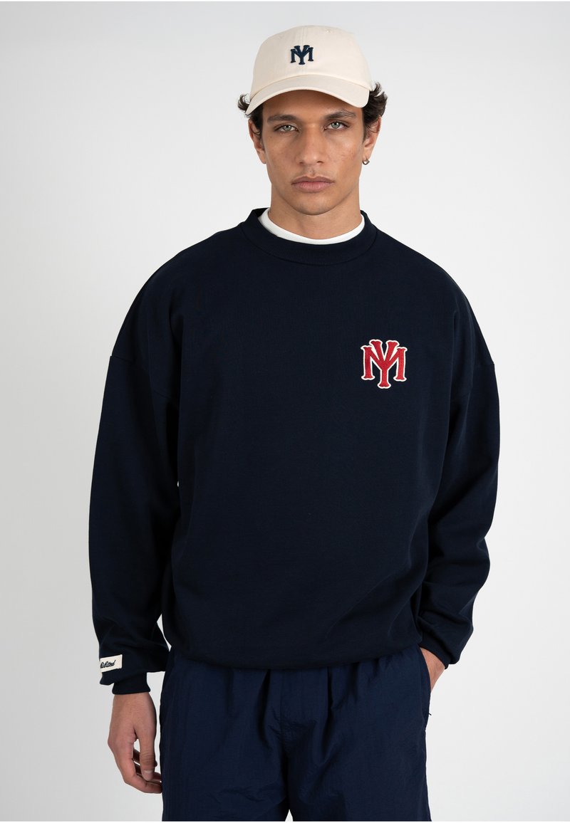 Jeune homme portant une casquette de baseball crème et un sweat-shirt bleu marine avec un logo "NY" rouge et blanc, debout avec une main dans la poche.
