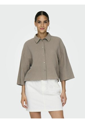 Cropped, loose-fitting, brown button-up shirt with a collar and textured fabric. Paired with a white skirt. Circular hoop earrings visible.