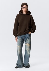 Felpa marrone con tasca frontale, abbinata a jeans in denim a gamba larga scoloriti e strappati. Stivali neri completano l'outfit.