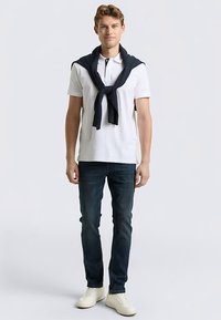 Polo blanc avec un pull marine noué autour des épaules, associé à un jean denim foncé et des baskets blanches. Design simple avec des couleurs contrastées.