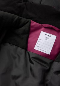 Musta ja harmaa takin sisäpuoli, jossa on viininpunainen paneeli ja valkoinen etiketti, jossa lukee koko 116 (5-6 vuotta) sekä viivat neljän nimen kirjoittamista varten.