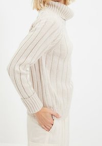 Woman wearing a cream ribbed turtleneck sweater with long sleeves, hand resting in pocket of light-colored pants, shown from side.