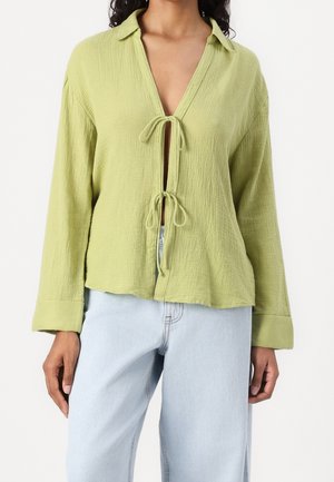 Jasnozielona, teksturowana bluzka z kołnierzykiem, długimi rękawami i związaną z przodu klamrą. W zestawieniu z jasno niebieskimi dżinsami.