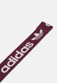 Maroon kangasta tehty hihna, jossa on valkoiset "adidas"-kirjaimet ja kolmonen raitalogo. Teksturoitu kudontamuoto, joka on litteä ja suorakulmainen.