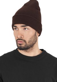 Brown knit beanie with a tapered top and ribbed texture. Worn with a black crew neck t-shirt, featuring a close-cropped hairstyle and beard.