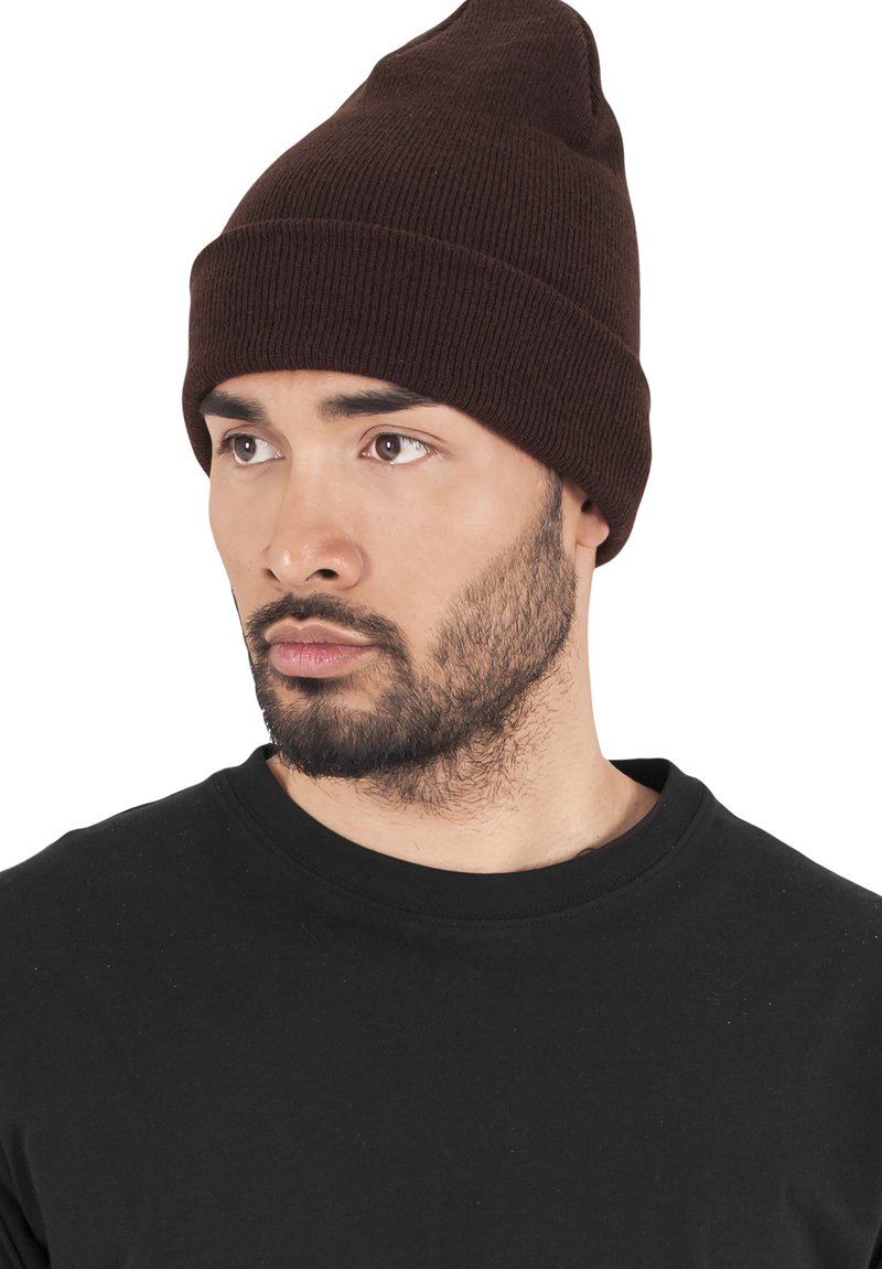 Brown knit beanie with a tapered top and ribbed texture. Worn with a black crew neck t-shirt, featuring a close-cropped hairstyle and beard.