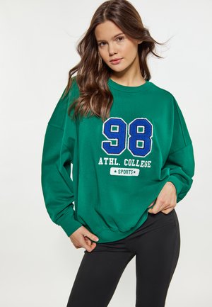 Jeune femme aux longs cheveux bruns porte un sweat-shirt vert avec l'inscription « 98 ATHL. COLLEGE SPORTS » et des leggings noirs, posant sur un fond blanc.