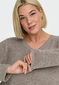 Blondýna upravující rukáv taupe pleteného svetru, nosící zlaté kruhové náušnice, s neutrálním výrazem na světle pozadí.