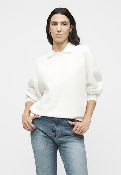 Femme aux cheveux foncés portant un pull blanc à manches longues avec col et un jean bleu, debout les mains près des poches devant un fond uni.