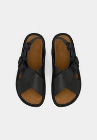 Μαύρα σανδάλια με σταυρωτούς ιμάντες από ματ συνθετικό υλικό, με ρυθμιζόμενες αγκράφες και καφέ εσωτερική σόλα με υφή.