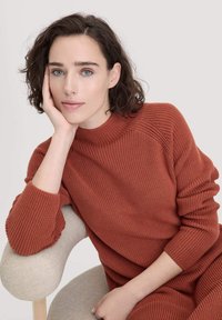 Gestrickter, rostfarbener Pullover mit einer gerippten Textur, ausgestattet mit einem hohen Kragen und lässigem Schnitt, kombiniert mit einem weichen, beigefarbenen Polstersessel.