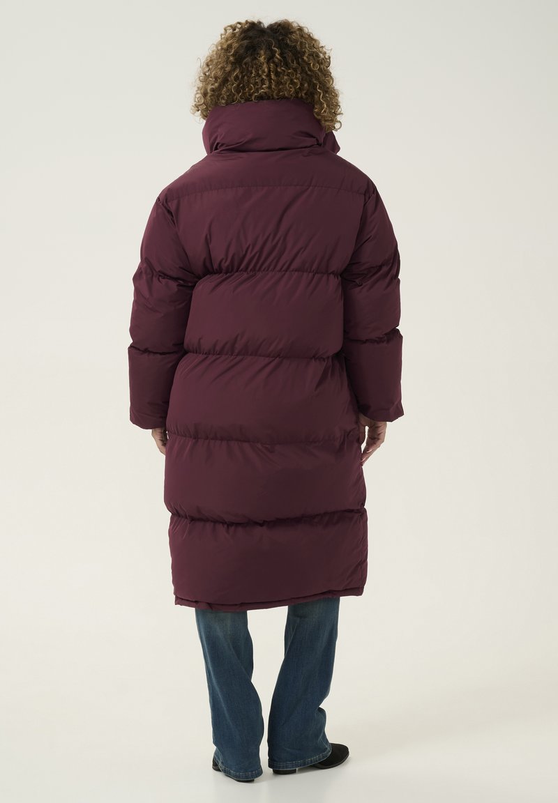 Lange bordeaux pufferjas met hoge kraag, voorzien van gestikte secties en lange mouwen. Draagt over blauwe jeans en zwarte schoenen.