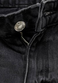 Črni denim tkanina z kovinsko gumbom in zadrgo. Ima vidne šive in rahlo teksturo, kar poudari trpežnost tkanine.