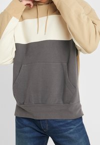 Sudadera con capucha con bloques de color beige, crema y gris oscuro. Cuenta con un bolsillo delantero y una capucha con cordón. Fabricada con tela suave.