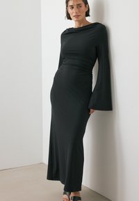 Svart långärmad klänning med figursydd överdel, draperad halsringning och utsvängda ärmar, tillverkad av ett glatt, elastiskt tyg. Enkel och elegant design.