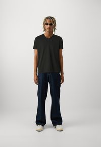 Melnā V-veida T-krekls, brīva piegriezuma, kopā ar vaļīgām zilo džinsu biksēm. Modelis valkā gludas saulesbrilles un baltas kedas. Vienkārša fona.